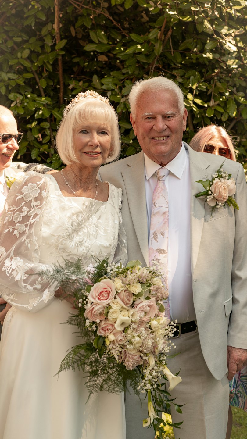 An older bride and groom smile while standing together outdoors.