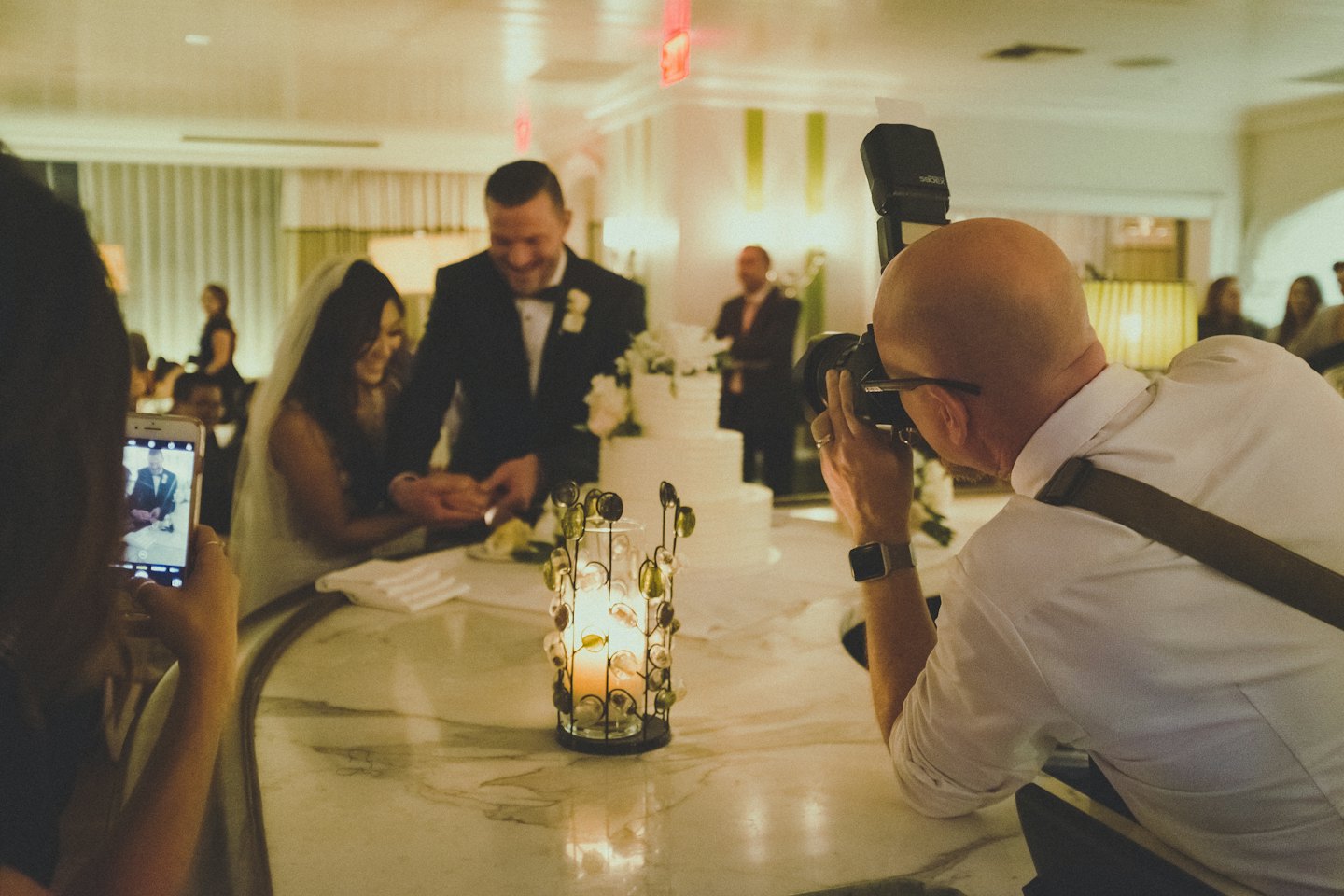 Bride and groom cut wedding cake while guest takes photo.