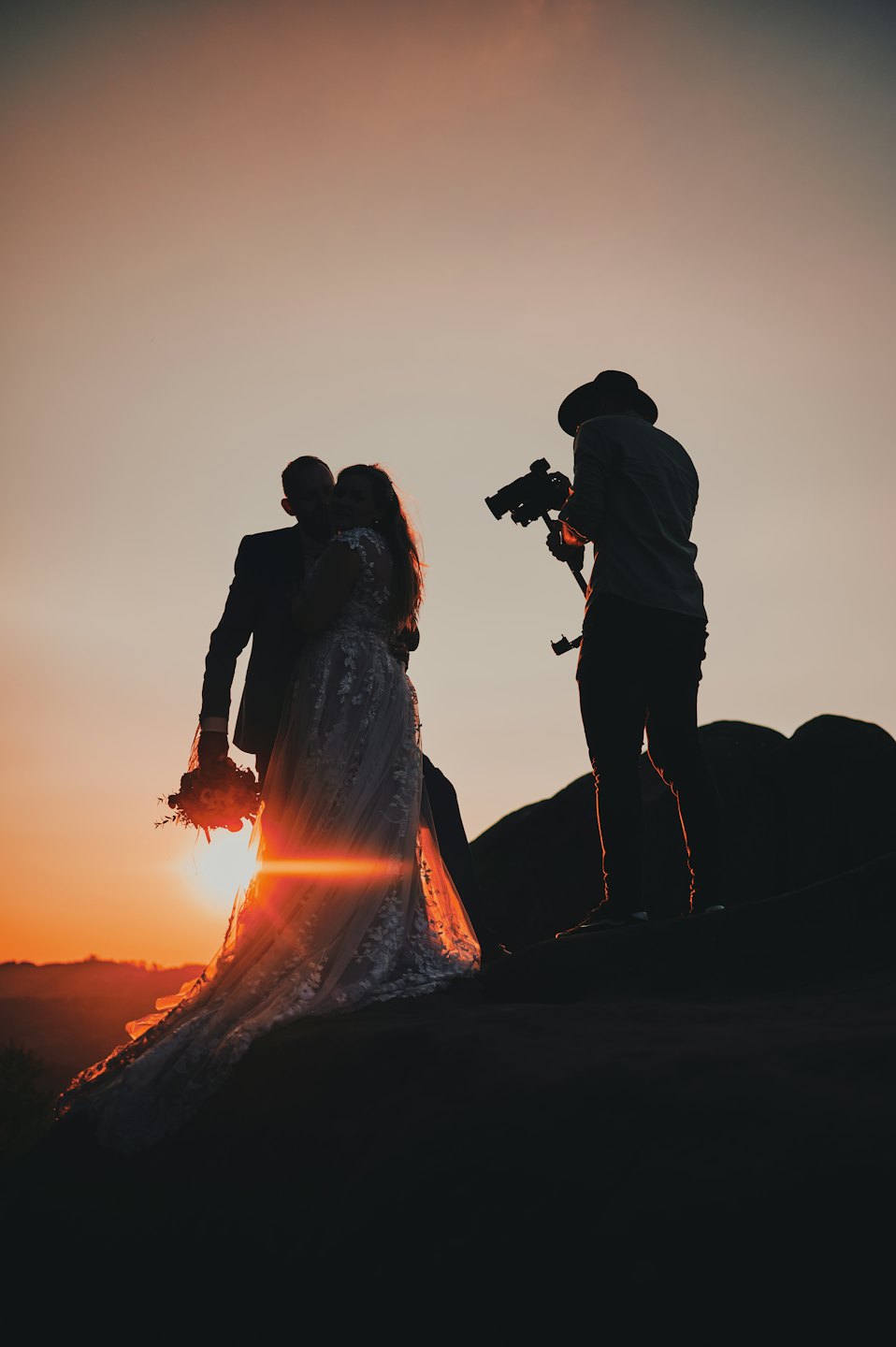 Newlyweds embrace on a rocky outcrop at sunset.