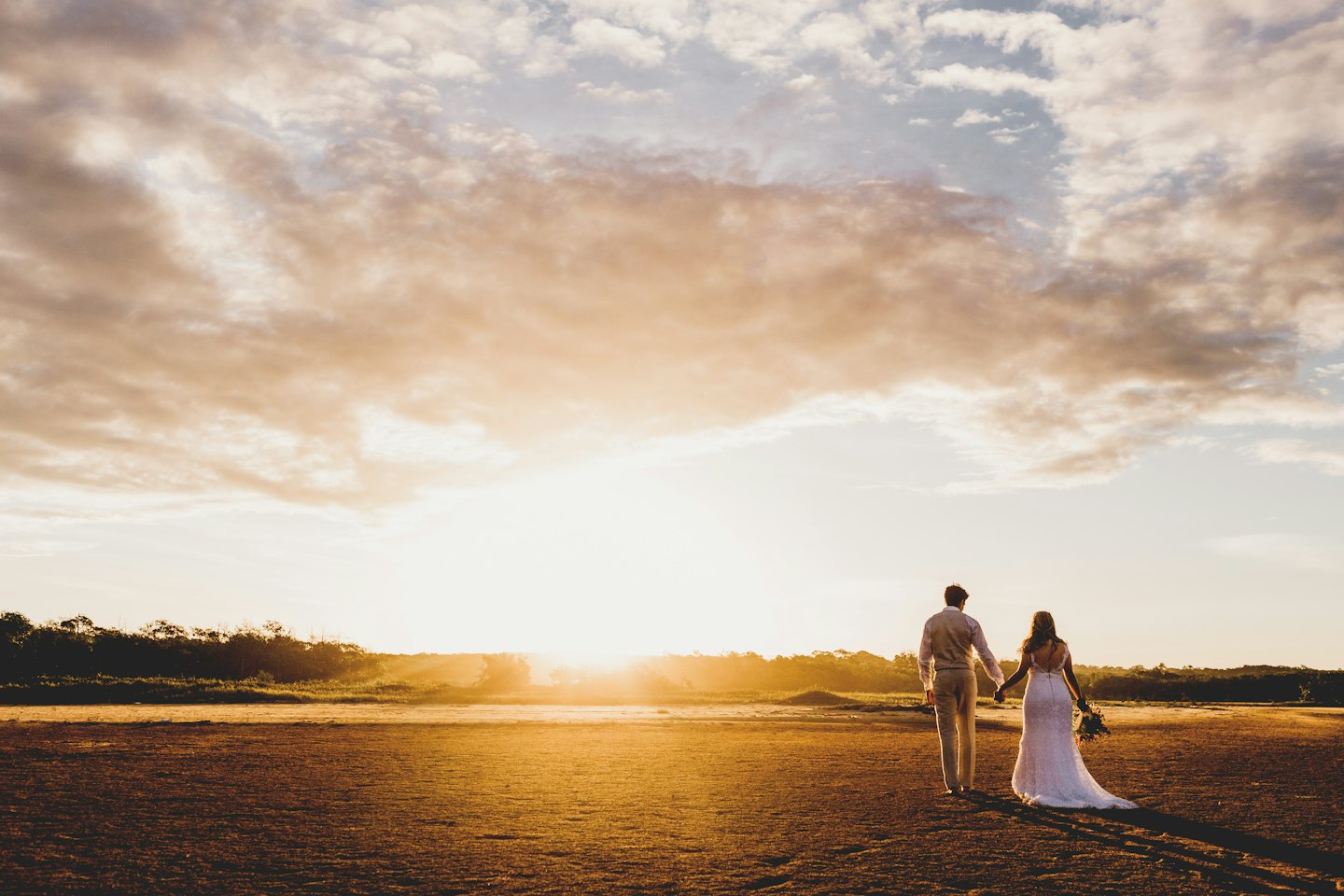 Newlyweds in wedding attire walk hand in hand across a field.